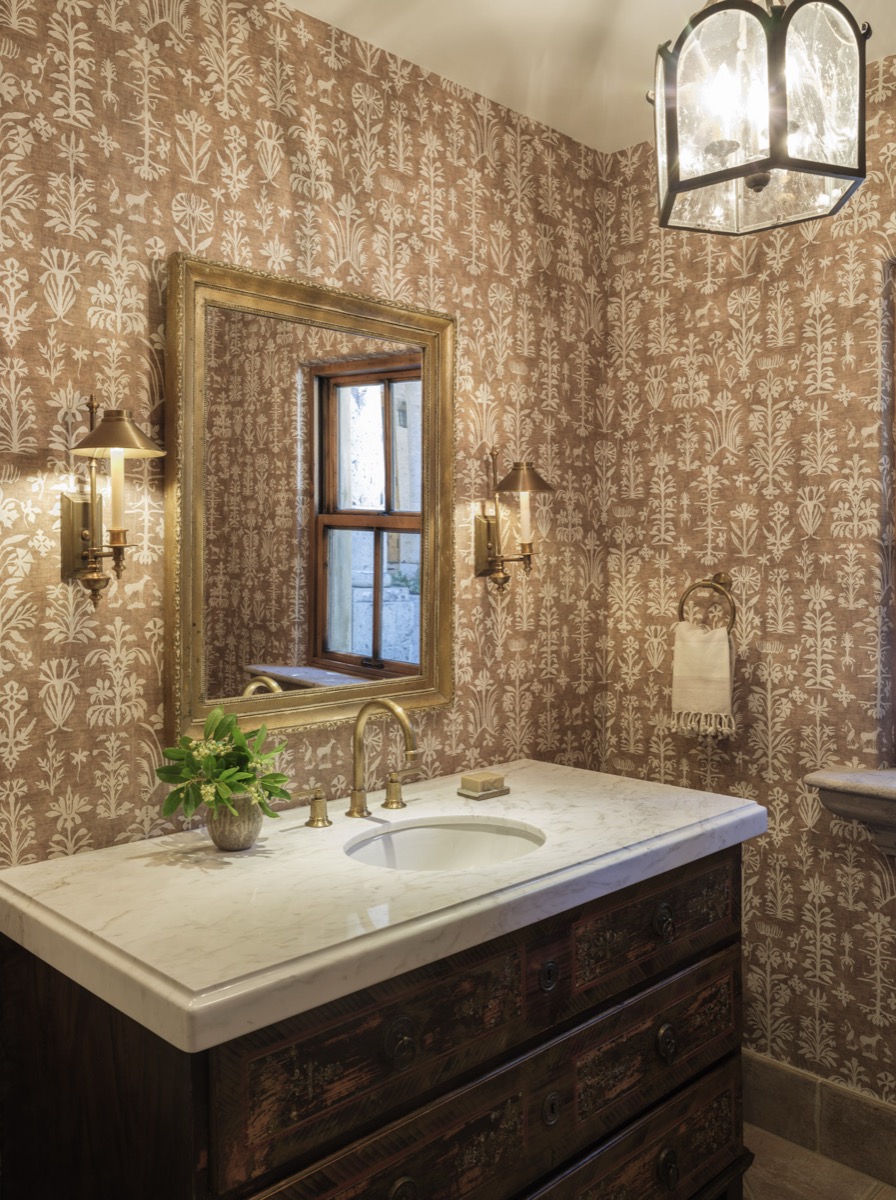 Powder room with patterned wallpaper, a wood vanity and brass sconces