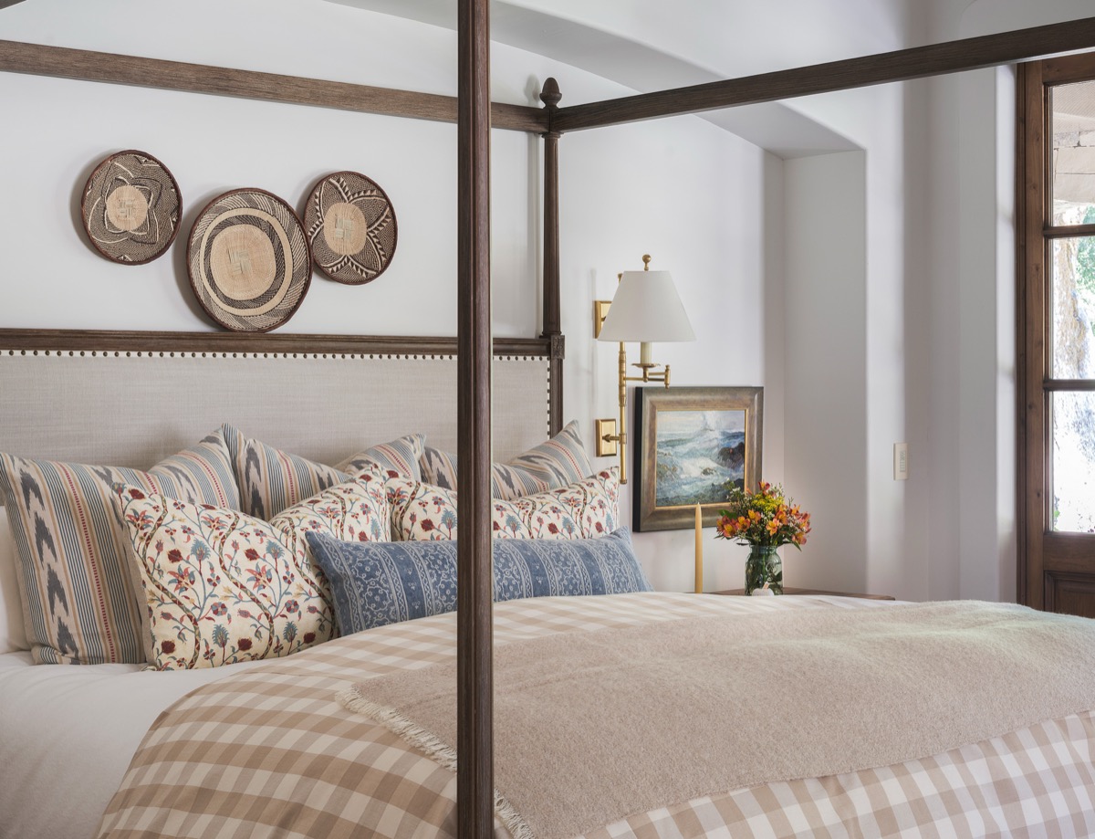 Four-poster bed dressed with layered patterned pillows and woven wall baskets